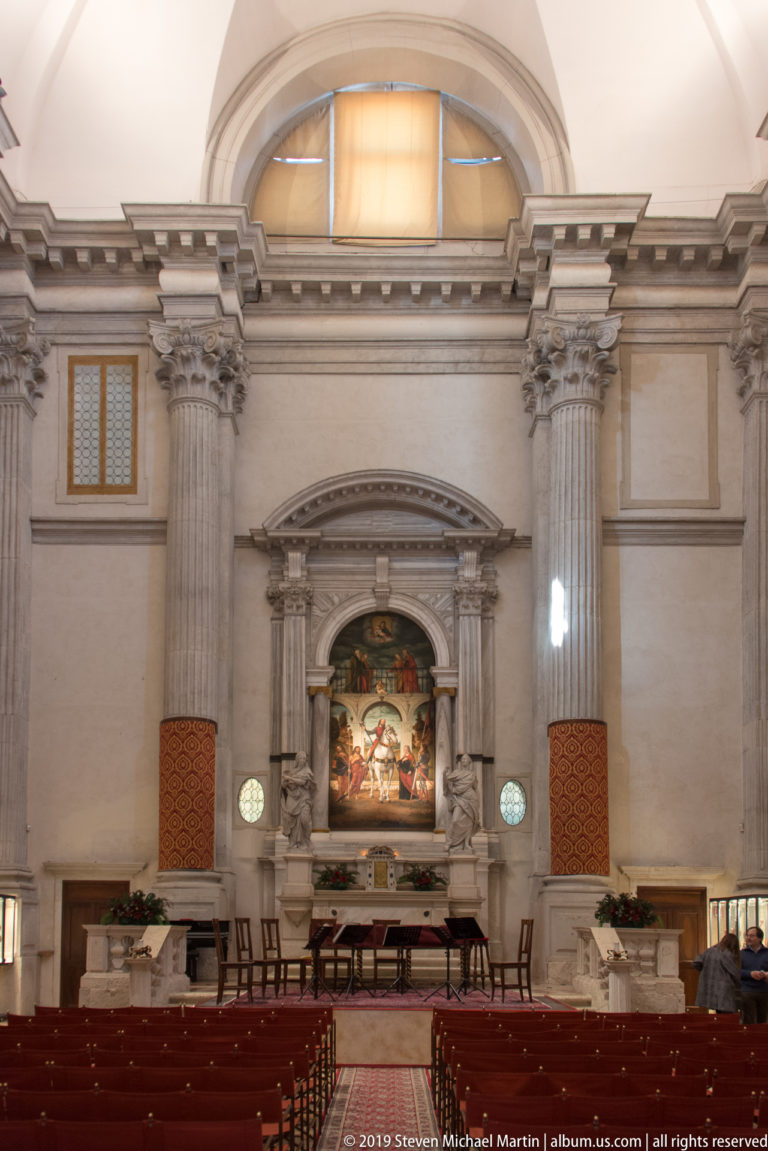 Wide church interior with rows of red chairs facing a side altar, monumental columns, and a large religious painting.