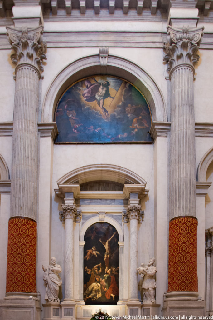 Side altar between tall fluted columns, with a dark altarpiece, sculpted figures, and a large arched painting above.