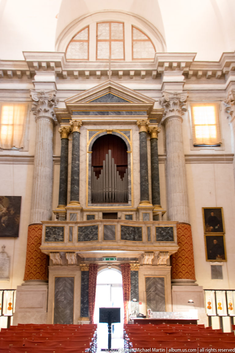 Pipe organ in a raised gallery framed by fluted columns and a triangular pediment above rows of red church seating.