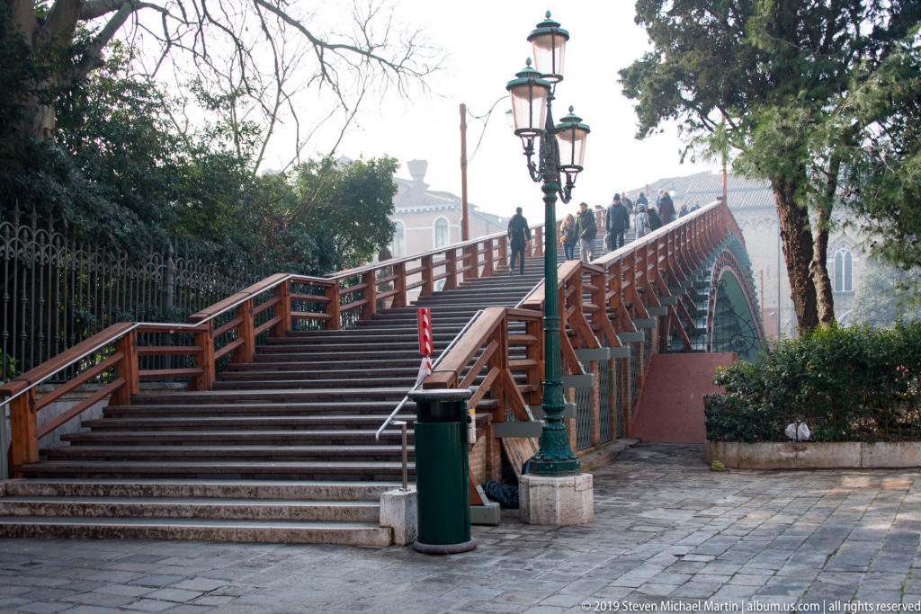 Wide steps rise onto the wooden Ponte dell'Accademia, with pedestrians, decorative lamps, railings, and trees along the approach.