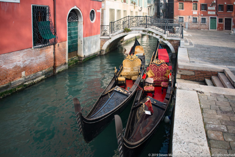 Decorated gondolas moored beside stone steps on a narrow canal, facing a small arched pedestrian bridge.