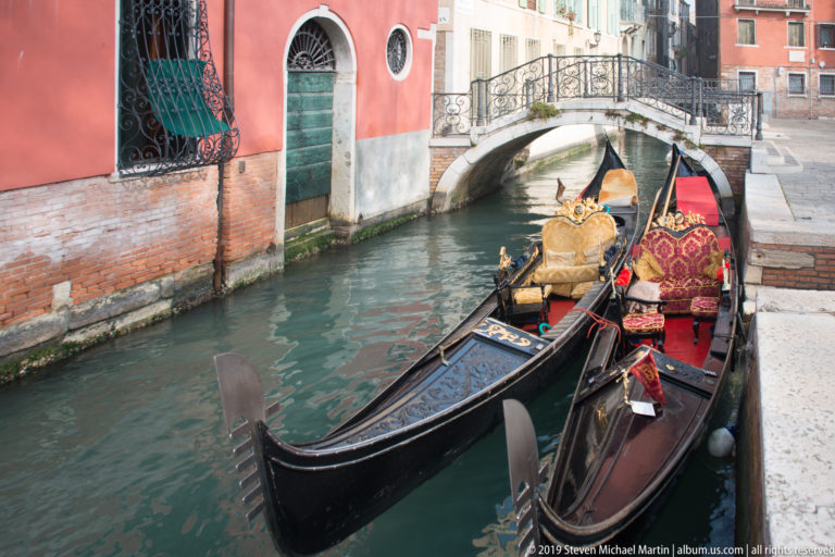 Two ornate black gondolas with red-and-gold seats moored beside a narrow canal, with a small arched bridge behind them.