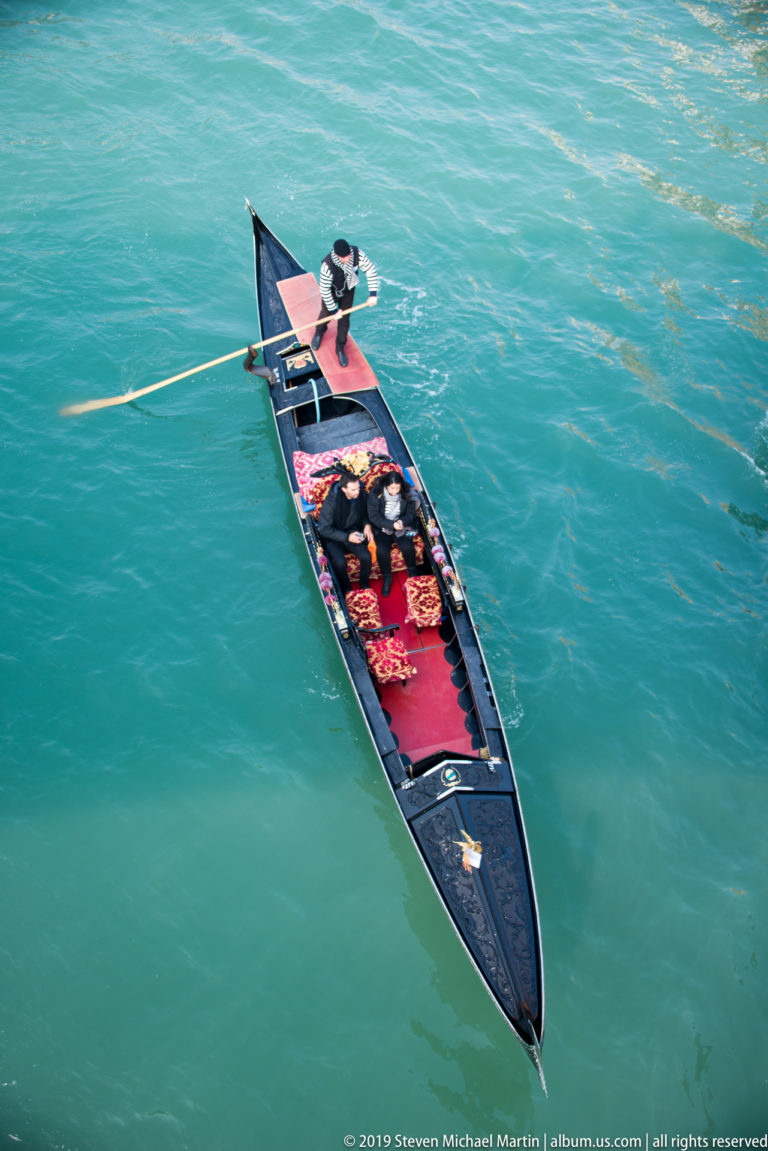 Black gondola seen from above on turquoise water, with red seats, a standing gondolier, and a long oar.