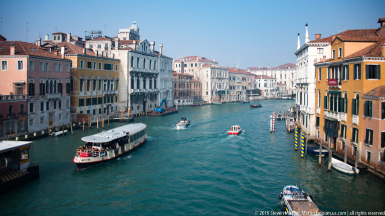 Elevated view along the Grand Canal with a vaporetto, small boats, palazzi, and striped mooring posts.