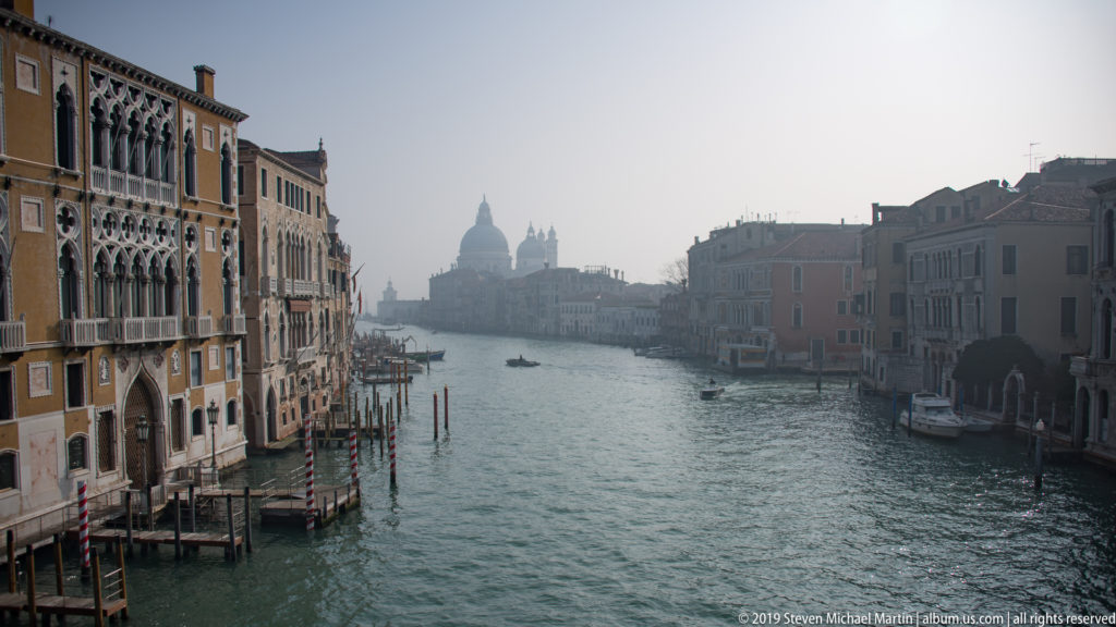 Backlit Grand Canal lined by palazzi, with small boats on the water and domes of Santa Maria della Salute in the hazy distance.