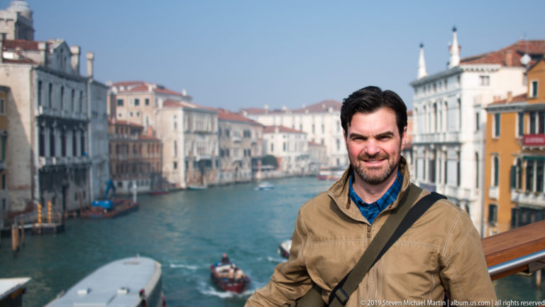 Close portrait of a man in a tan jacket at a wooden bridge railing, with the Grand Canal softly blurred behind him.