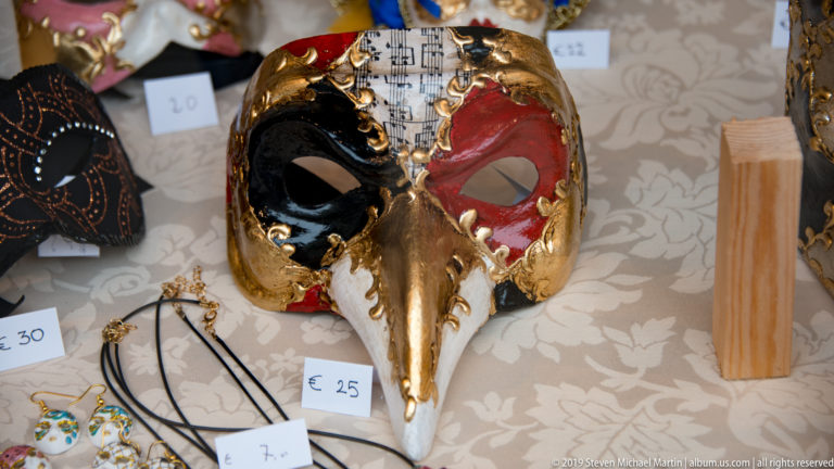 Gold long-beaked Venetian mask with red and black eye decoration displayed among jewelry and other masks with euro price tags.