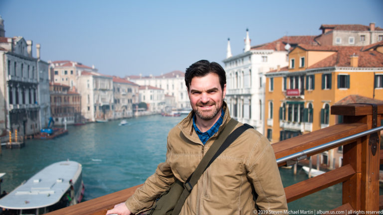 Man in a tan jacket leans on a wooden railing above the Grand Canal, with a vaporetto and palazzi behind him.