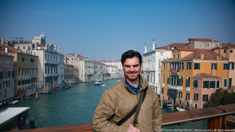 Man in a tan jacket stands at a wooden railing with the Grand Canal, boats, and colorful Venetian palazzi behind him.