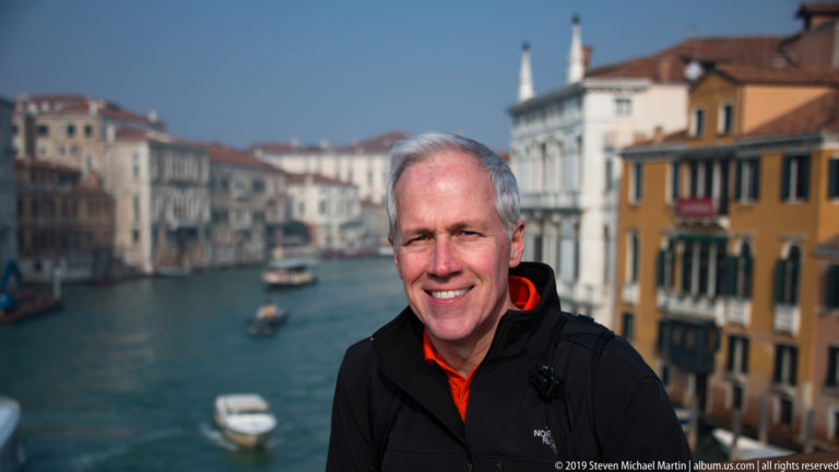 Smiling gray-haired man in a dark jacket stands above the Grand Canal with boats and colorful palazzi blurred behind him.