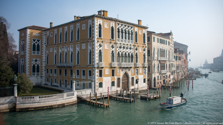 Ornate ocher palace with pointed Gothic-style windows and balconies beside the Grand Canal as a small motorboat passes.