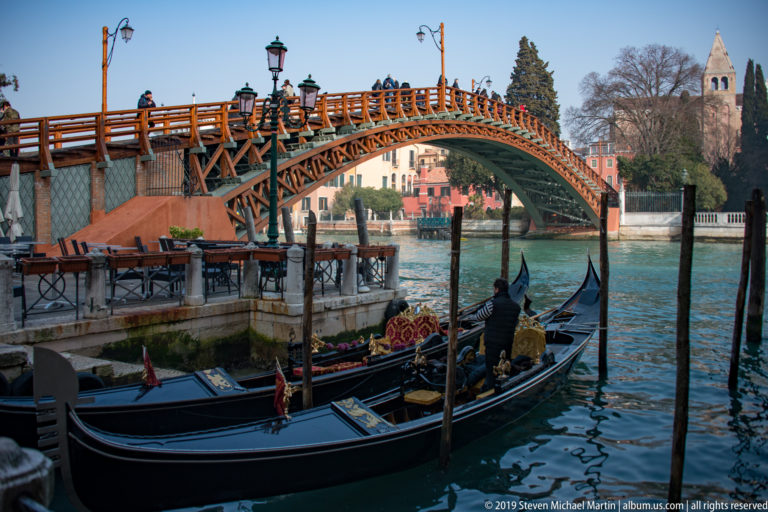 Wooden Ponte dell'Accademia arches over the Grand Canal above moored black gondolas and a gondolier at a waterside landing.