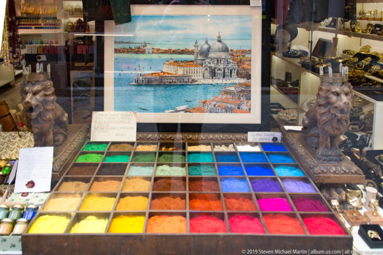 Shop window with a grid of brightly colored pigment trays, decorative objects, and a framed view of Santa Maria della Salute.