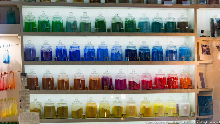 Shelves of glass jars filled with vivid green, blue, brown, red, orange, and yellow powdered pigments.