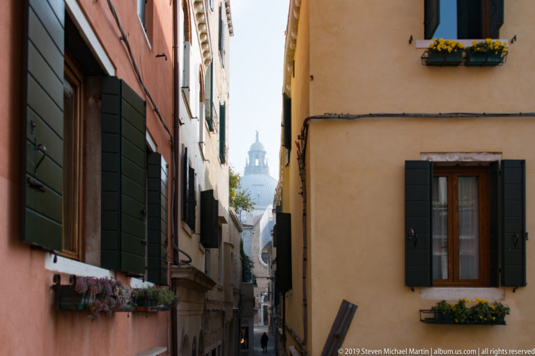 Narrow Venetian alley between peach and ochre buildings with dark shutters, flower boxes, and a pale dome visible at the far end.
