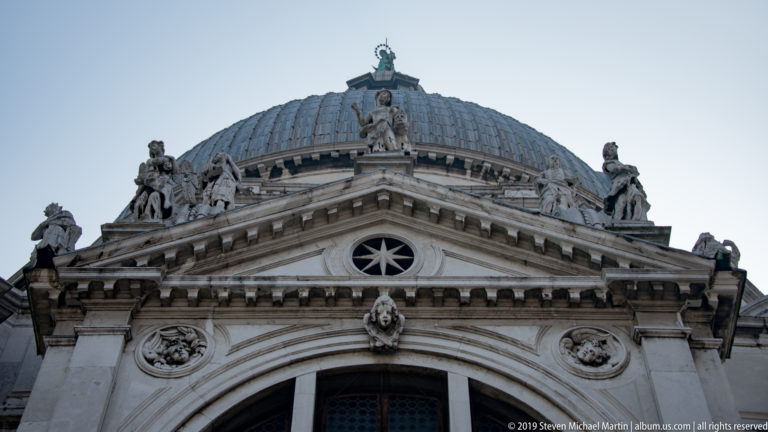 Upper façade of Santa Maria della Salute with triangular pediment, round star-like window, statues, and dome behind.