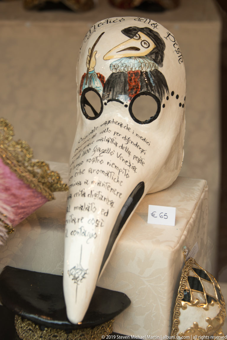 Cream long-beaked Venetian mask decorated with script and painted motifs, displayed with a small price card in a shop.