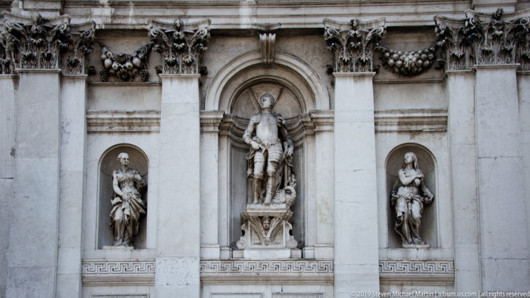 Three stone statues set in niches between Corinthian pilasters, carved garlands, and a Greek-key frieze.