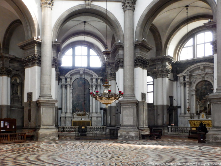 Side of a grand church interior with stone piers, high arches, altar, hanging lamp, and patterned marble floor.