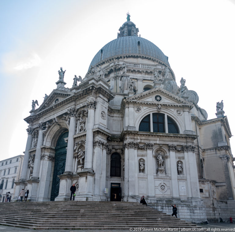 Low-angle exterior of Santa Maria della Salute with monumental arched entrance, columns, statues, and large dome.
