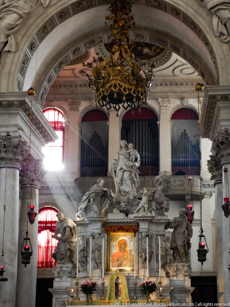 Marble sculptural ensemble and glowing icon beneath organ pipes, with red votive lamps and bright side light.