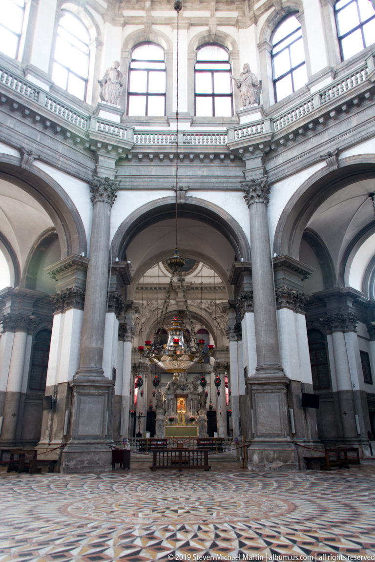 Wide church interior with patterned marble floor, massive stone piers, round arches, high windows, and an altar in the distance.