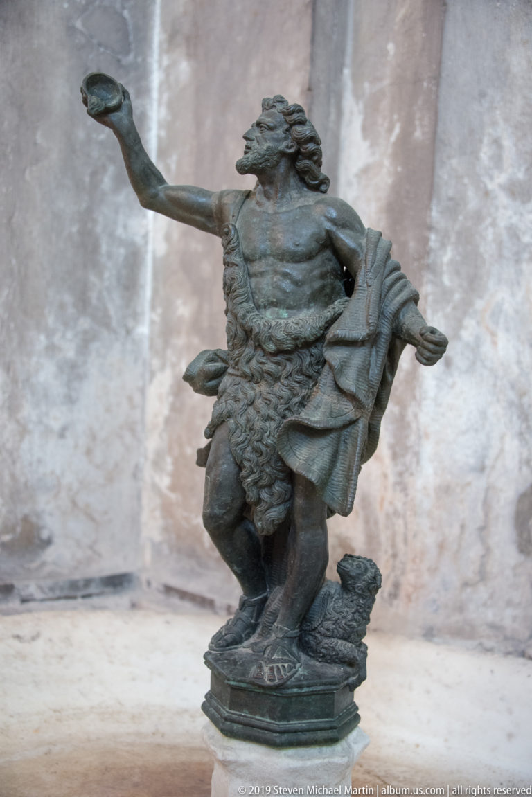 Small dark bronze statuette of a muscular male figure striding with one arm extended and drapery around his body.