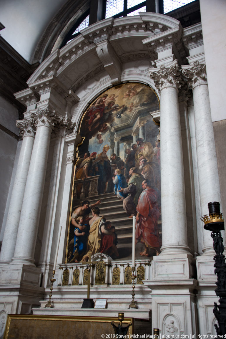 Large religious painting above a side altar, framed by paired pale columns and curved stone architecture.