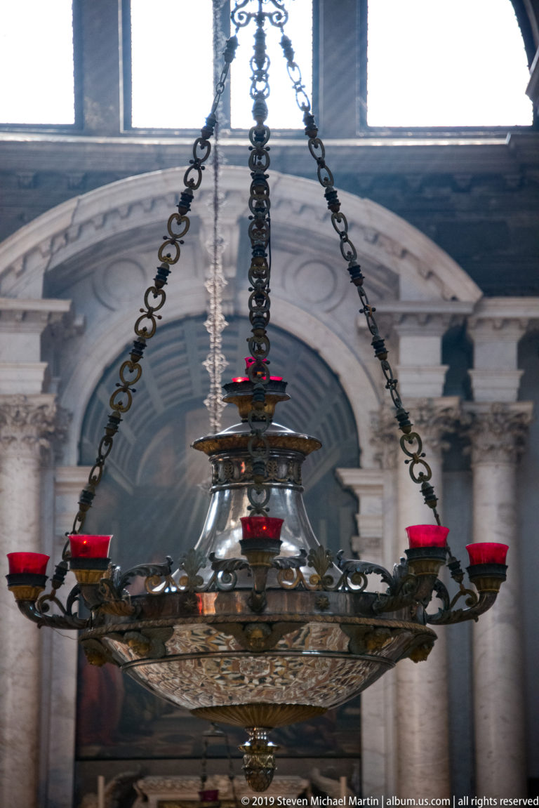 Wide view of an ornate hanging lamp on long chains before a pale church arch and high window.