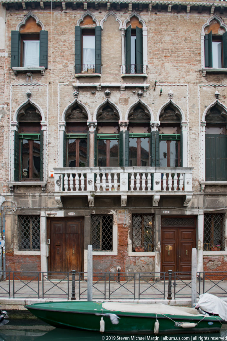 Weathered brick canal-side façade with pointed-arch windows, pale stone trim, balcony, green shutters, and wooden doors.