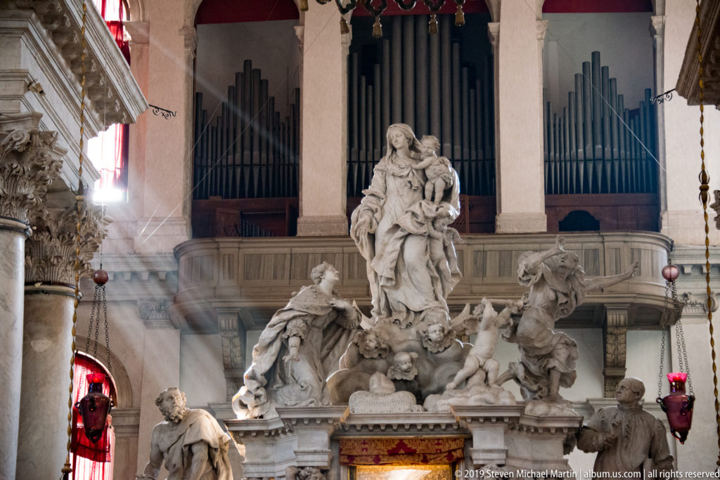 Large marble sculptural ensemble beneath organ pipes, with a painted icon below and red votive lamps at the sides.