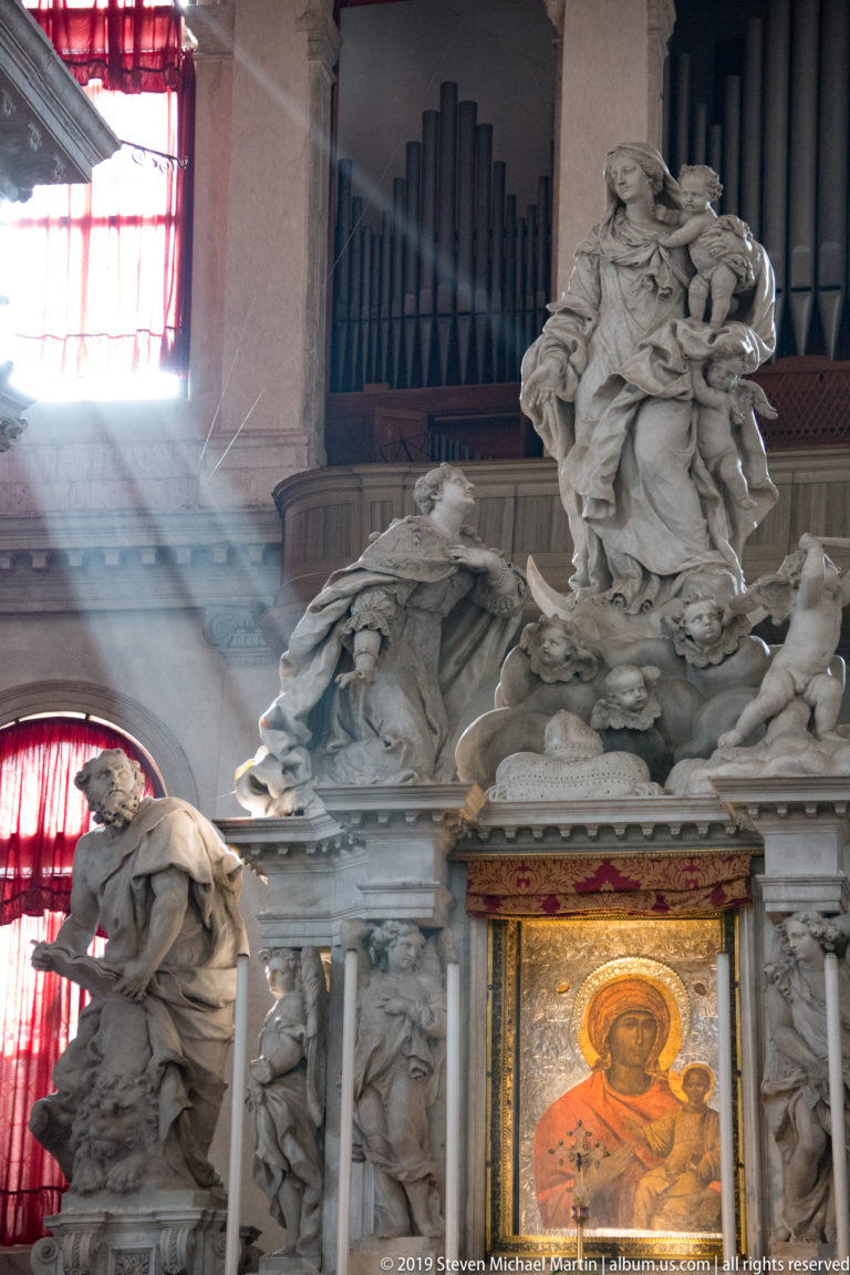 Painted icon beneath a large marble sculptural group, with organ pipes and a diagonal sunbeam behind.