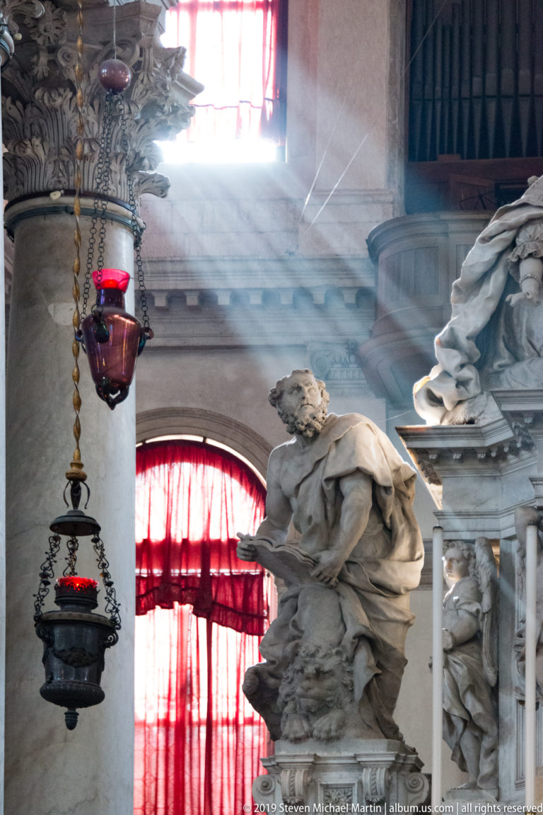 Sunbeam crossing marble church sculptures and red hanging votive lamps, with organ pipes and curtained windows behind.