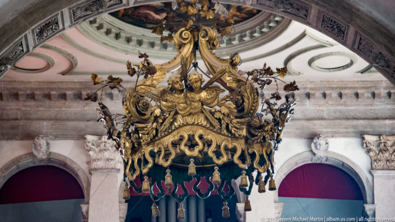 Ornate suspended church canopy with gilded scrollwork, blue-green fabric panels, tassels, and sculpted cresting.