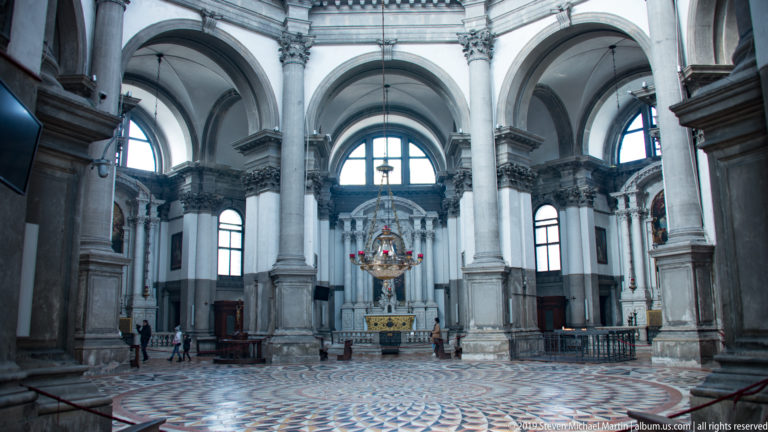 Spacious church interior with gray stone piers, high arches, patterned marble floor, tall windows, and an altar ahead.