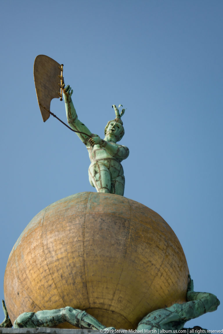 Low-angle view of Fortuna holding a sail atop a large gilded globe with sculpted figures at its base.
