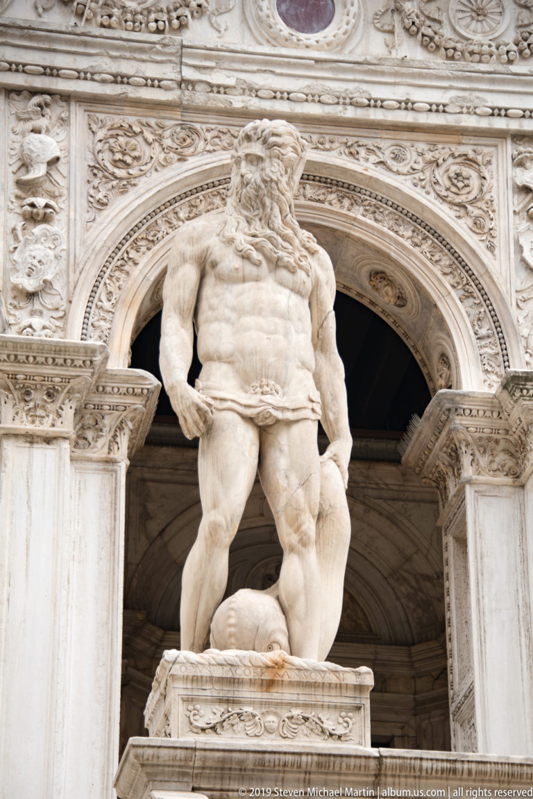 Bearded marble male statue stands in an ornate arched niche, one foot raised on a rounded sculptural support.