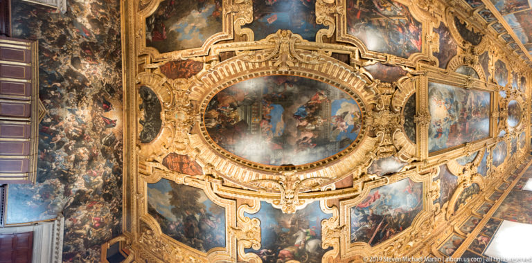 Oblique view of a richly gilded ceiling with an oval central painting and surrounding rectangular scenes.