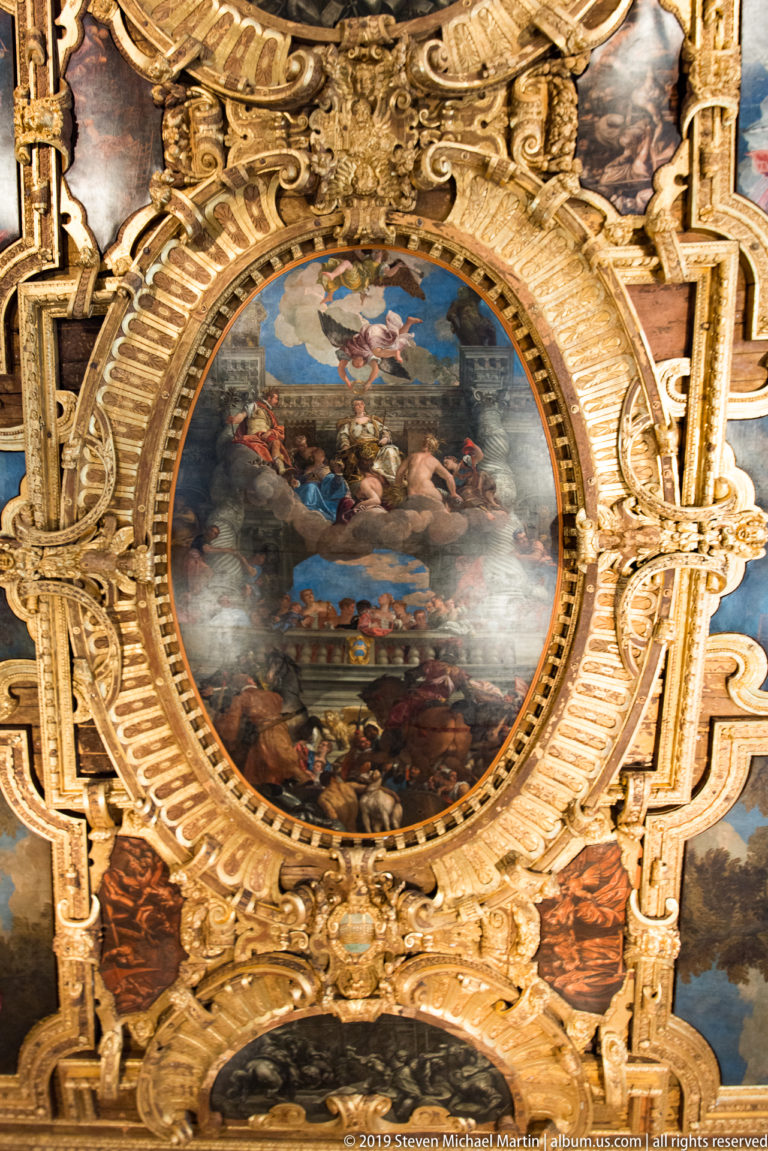 Oval narrative ceiling painting surrounded by elaborate gilded carved moldings and adjoining painted compartments.