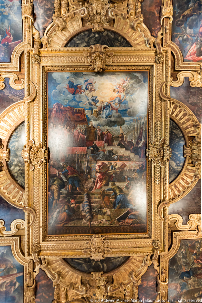 Rectangular ceiling painting of figures in a fortified landscape, surrounded by heavy gilded carving and partial oval side panels.