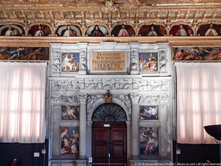Frontal view of the Francesco Morosini doorway beneath a painted portrait frieze, with curtains and wall paintings on both sides.