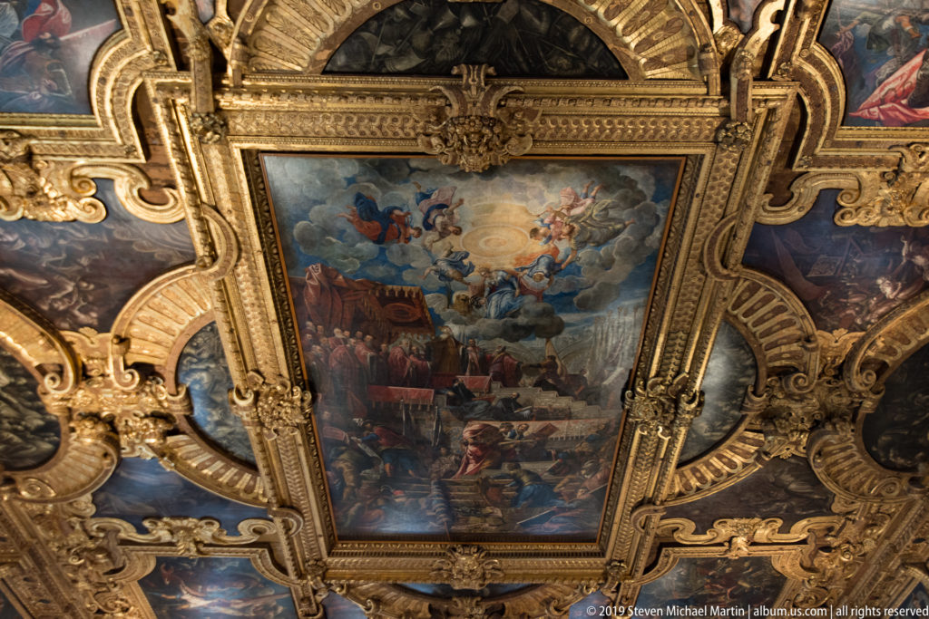 Large rectangular ceiling painting with figures and architecture below a blue sky, framed by heavy gilded carving and oval side panels.