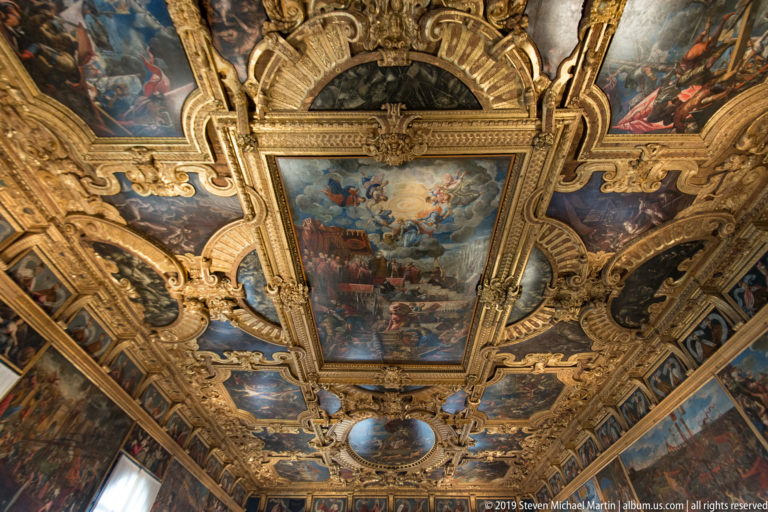 Upward view of a rectangular narrative ceiling painting surrounded by gilded scrollwork, smaller painted panels, and dark lunettes.