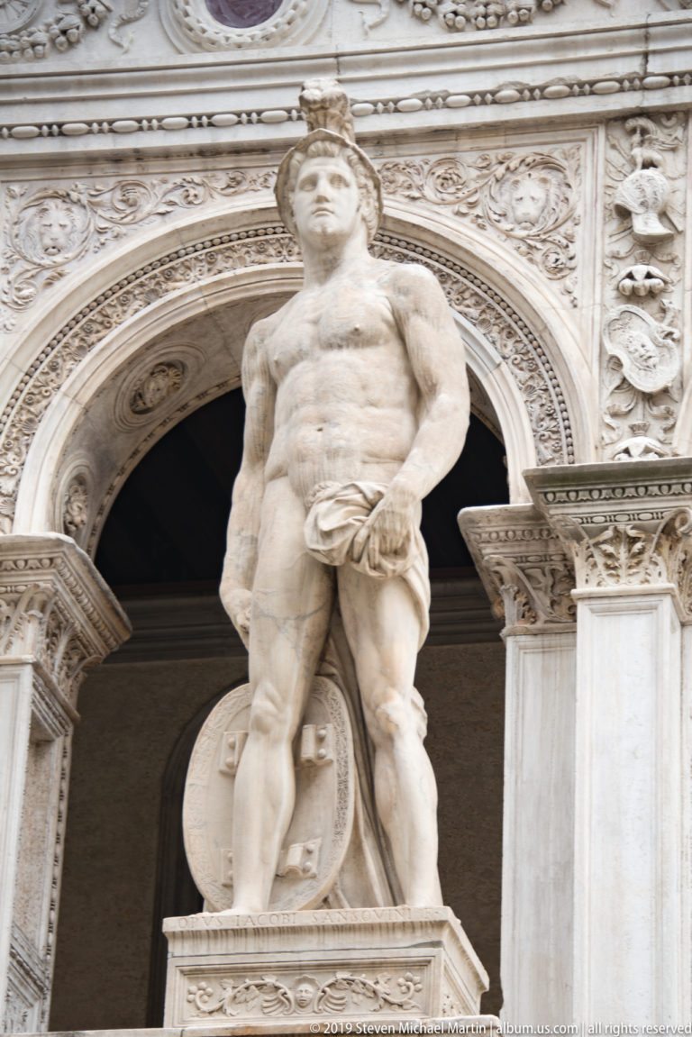 Standing marble male figure in front of an ornate round arch, with carved columns and relief ornament surrounding the niche.