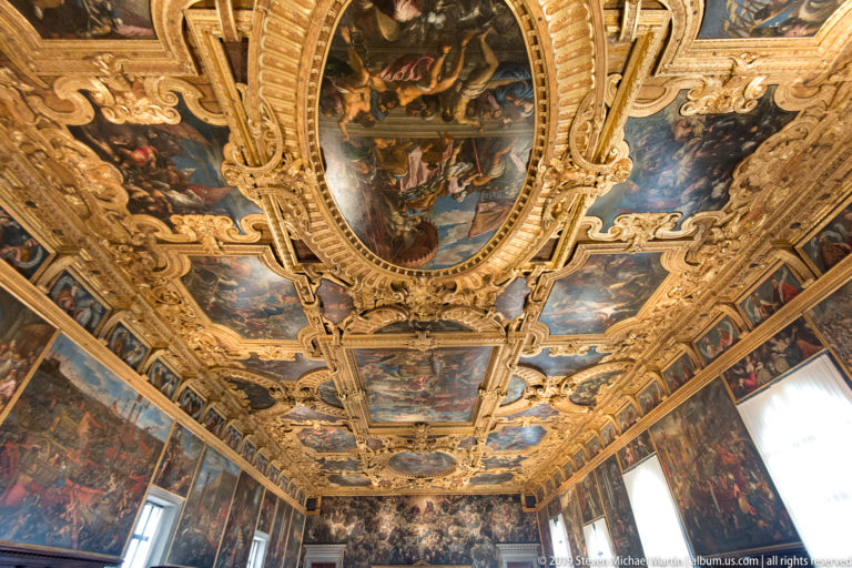 Long view through a grand hall with an elaborate gilded painted ceiling, large wall canvases, and tall bright windows.