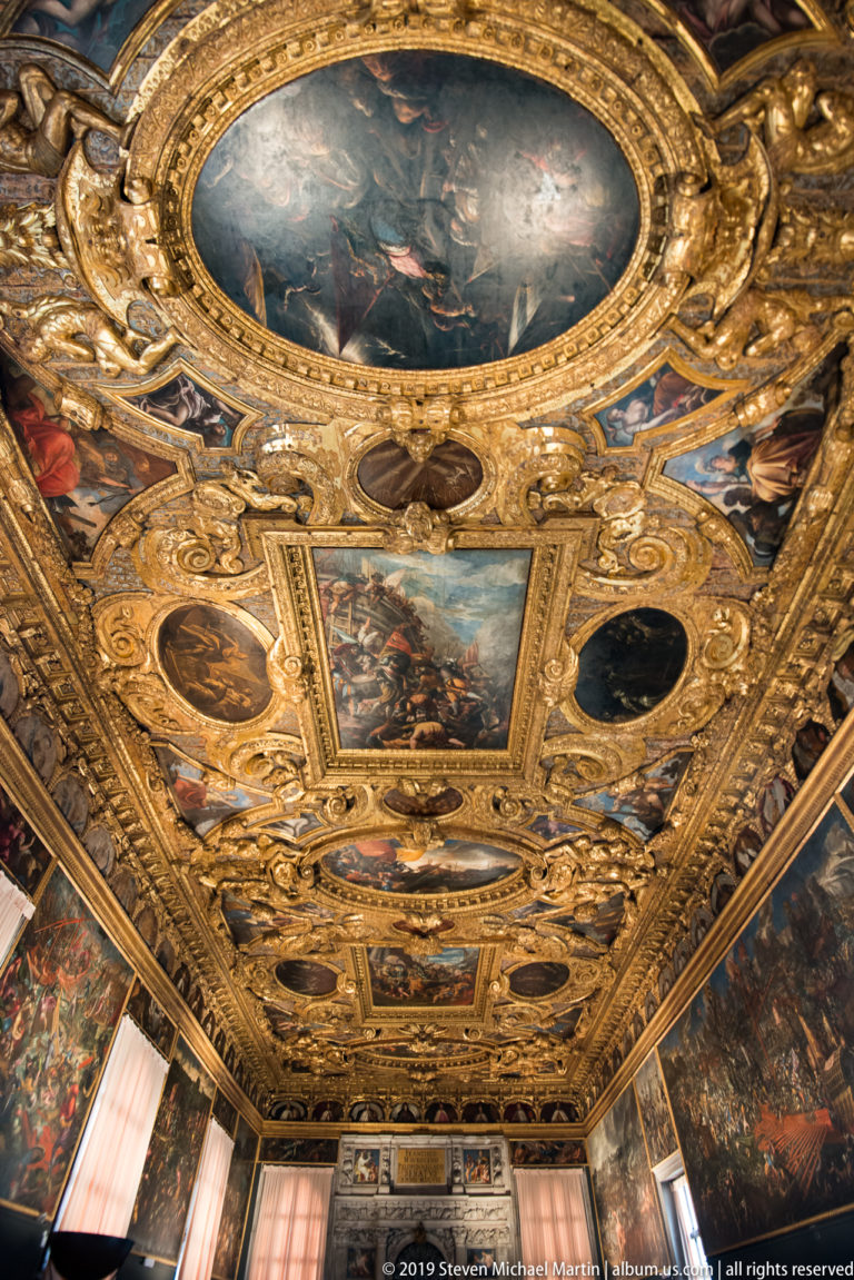 Oblique view across a richly gilded ceiling filled with oval and rectangular narrative paintings in carved frames.
