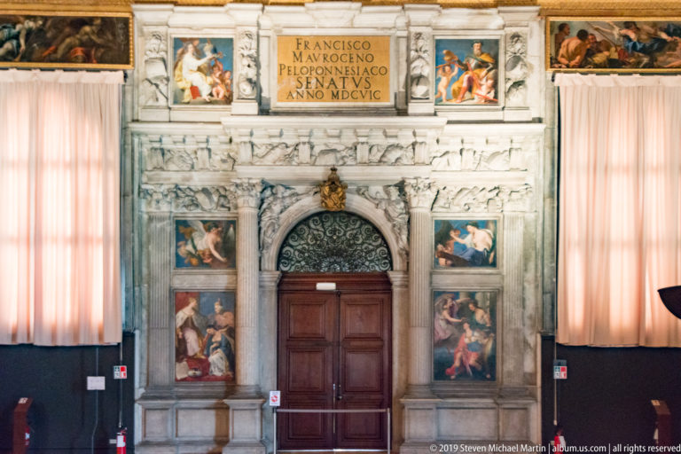 Ornate marble doorway with a Latin inscription naming Francesco Morosini, painted panels, relief sculpture, and a dark wooden door.