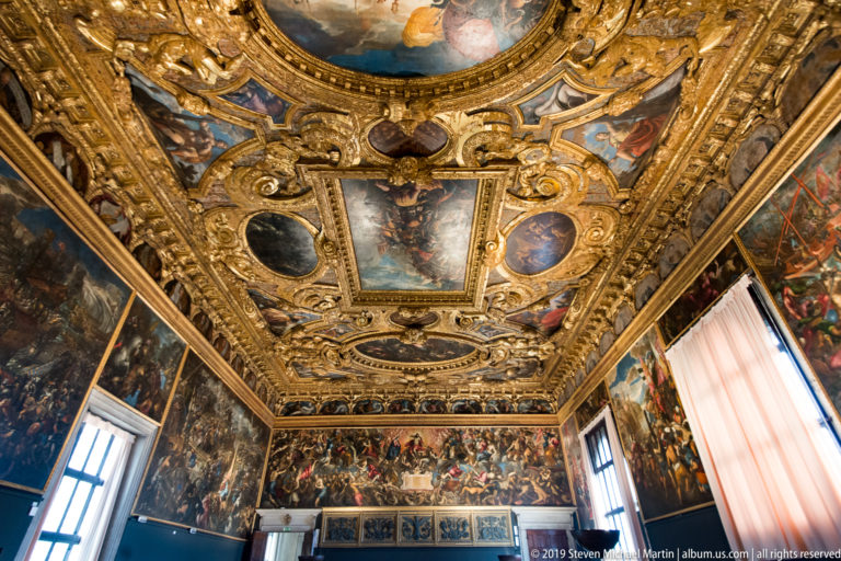 Wide interior view of a gilded coffered ceiling with many painted compartments above walls covered in narrative paintings.