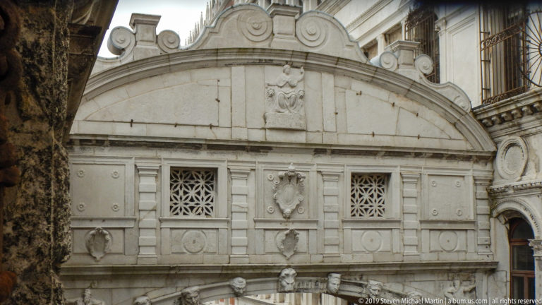 Close view of the Bridge of Sighs with paired lattice windows, carved shields, pilasters, and a curved stone pediment.