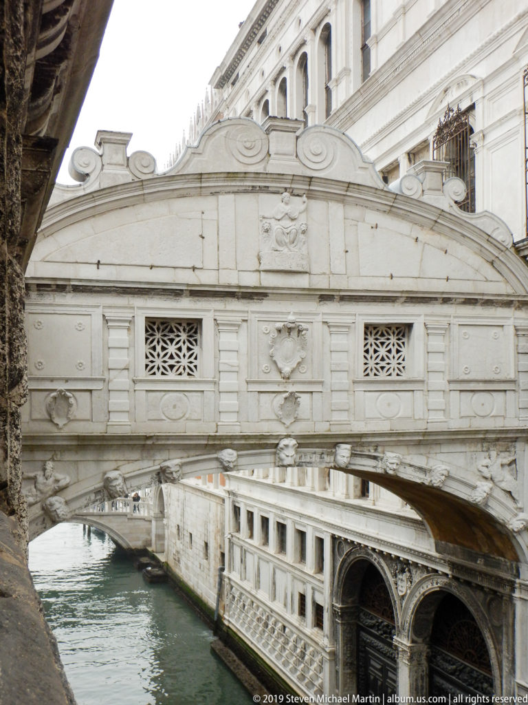 Pale stone Bridge of Sighs crossing a narrow canal, with lattice windows, relief ornament, and an enclosed passage.